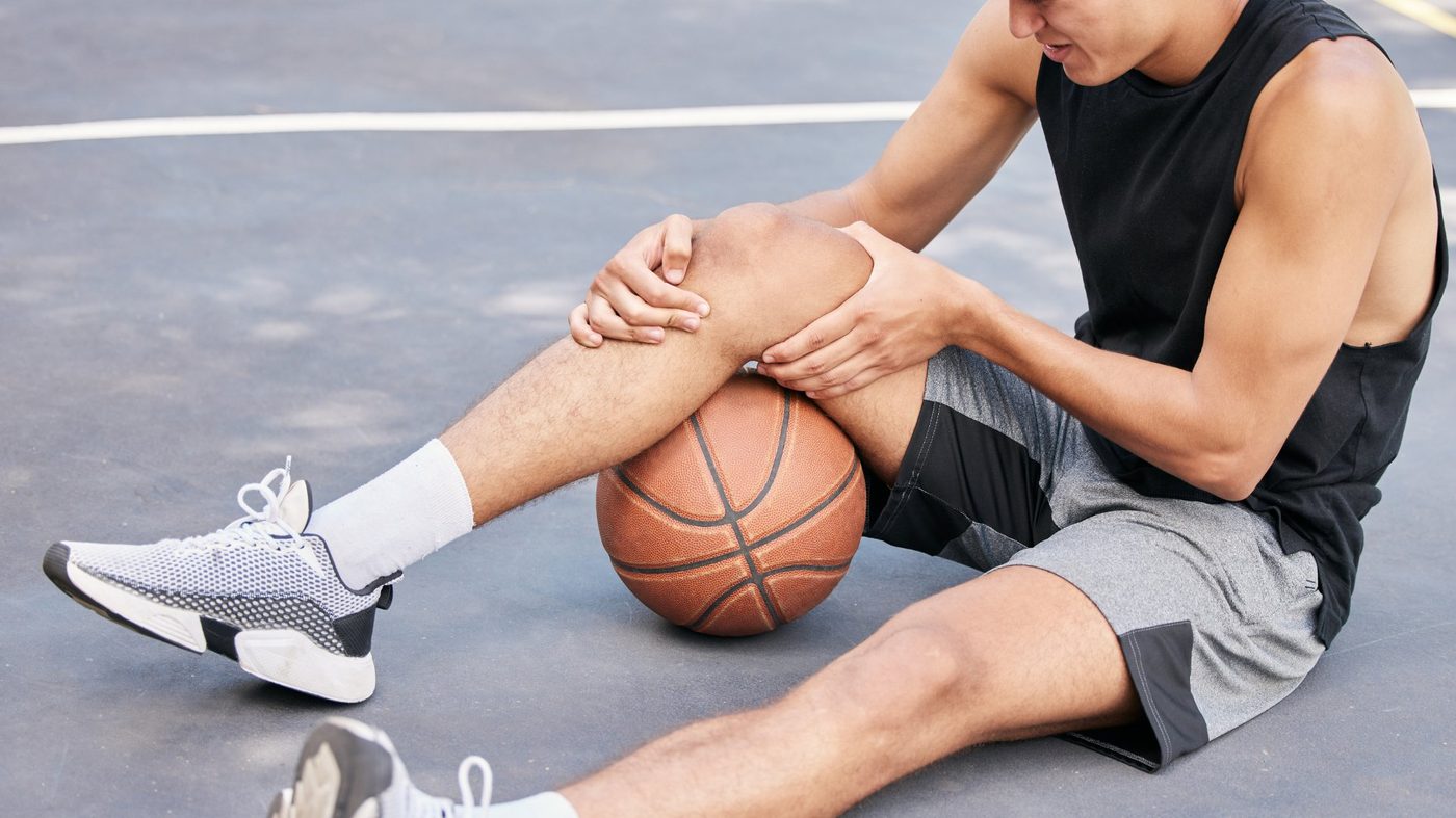 Young athlete sitting on a basketball court holding his knee after a sports injury